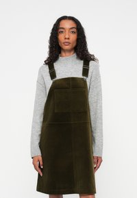 Nainen, jolla on kihara hius, pukeutunut oliivinvihreään samettipinnalliseen pinafore-mekkoon, jonka alla on vaalea harmaa neulepusero, seisoo yksinkertaista taustaa vasten.