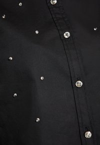 Černá košile s knoflíky, která má hladkou texturu, rozptýlené ozdoby z rhinestones a klasické detaily zapínání na knoflíky.