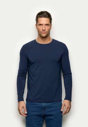 Mann mit kurzen dunklen Haaren, der ein dunkelblaues Langarmhemd und blaue Jeans trägt, vor einem schlichten hellen Hintergrund stehend.