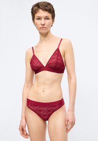 Γυναίκα με κοντά μαλλιά που φοράει κόκκινο δαντελένιο σουτιέν και ταιριαστά δαντελένια εσώρουχα, στέκεται μπροστά σε απλό φωτεινό φόντο.