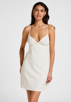 Jonge vrouw met lang donker haar draagt een witte mouwloze V-halsjurk met dunne bandjes en een subtiel gestippeld patroon, staand tegen een effen achtergrond.