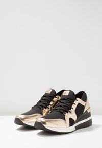 Czarno-złote buty sportowe z teksturowaną siateczkową cholewką, metalicznymi akcentami, czarnymi sznurówkami i podeszwą na klinie.