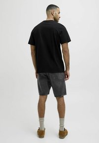 Camiseta de algodón negra vista desde la parte de atrás, combinada con shorts de mezclilla gris. Calcetines grises y zapatos marrones completan el conjunto. Diseño sencillo.