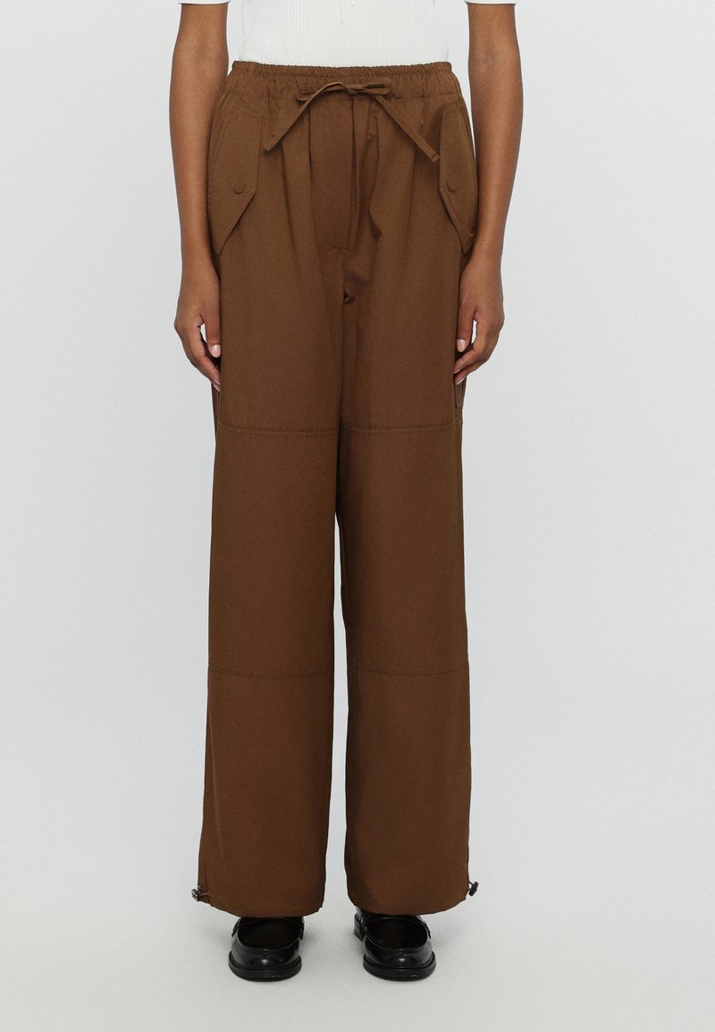 Brown wide-leg trousers with an elastic waistband, drawstring, side pockets, and zippered cuffs, made from a smooth, durable fabric.
