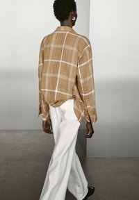 Langarm, geruit shirt in bruin en wit, gecombineerd met witte wijde broek. Het shirt heeft een ontspannen pasvorm en knoopdetails.