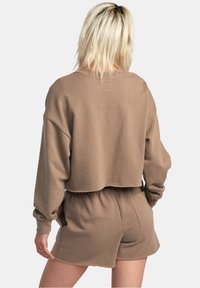 Felpa corta marrone con maniche lunghe, realizzata in tessuto morbido, abbinata a pantaloni corti coordinati, dotati di vita elastica e tasca.
