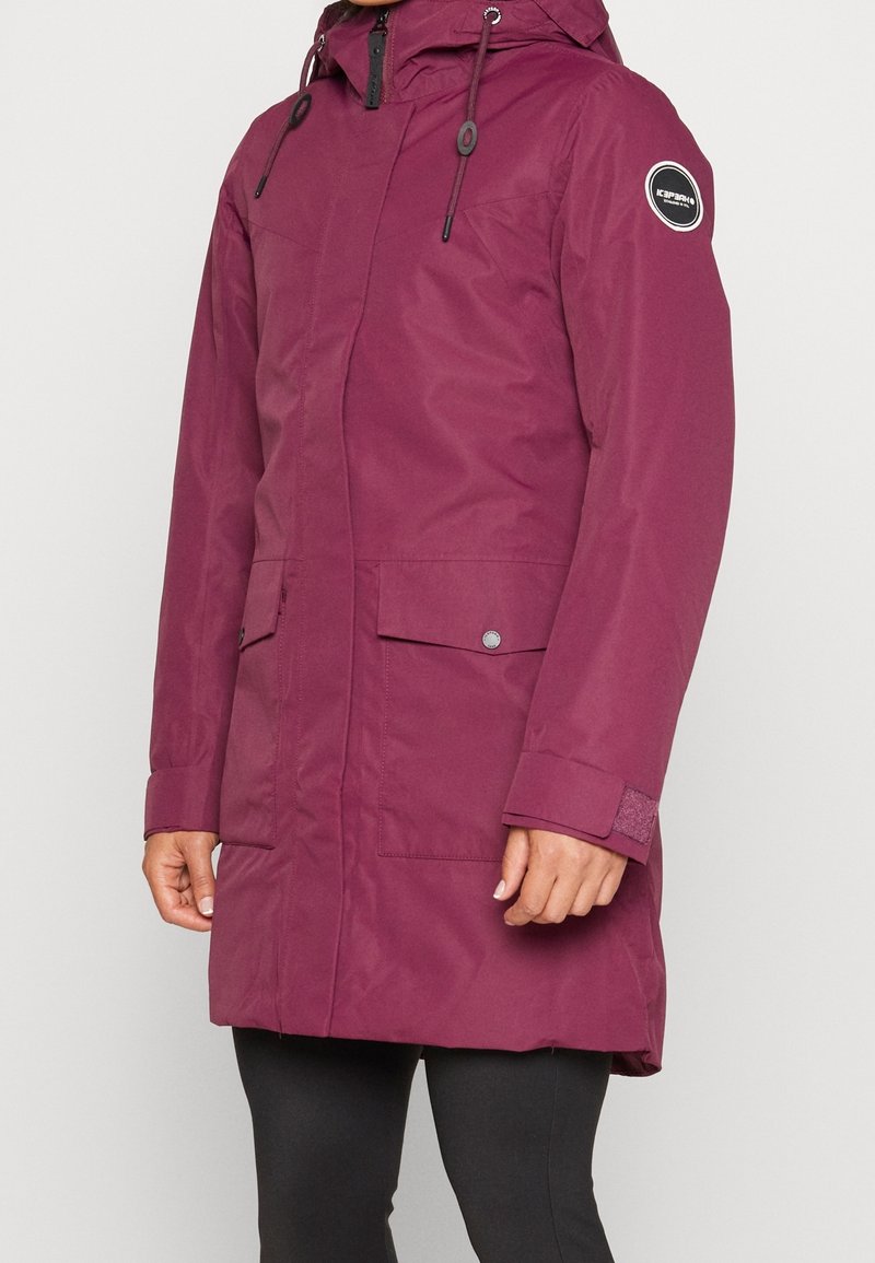 Manteau d'hiver violet mi-long à capuche avec poches avant, fermeture éclair et boutons-pression, porté sur des leggings noirs.