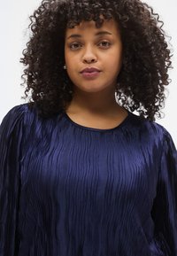 Μπλε μπλούζα σε ναυτικό μπλε χρώμα με πλισέ υφή, στρογγυλή λαιμόκοψη και μακριά μανίκια με ραφές που δημιουργούν εφέ συλλογής. Το ύφασμα έχει μεταξένιο φινίρισμα.