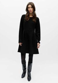 Robe noire à manches longues avec un corsage ajusté et une jupe évasée. Caractérisée par une texture douce et un col rond. Assortie à des bottes noires à motifs.