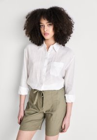 Camisa de lino blanca con botones y un bolsillo en el pecho, combinada con pantalones cortos verde oliva que cuentan con una cintura con cordón y una textura suave.