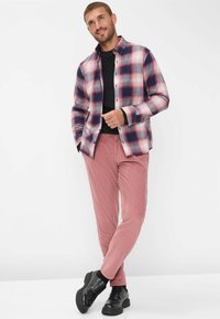 Camicia di flanella da uomo a quadri rossi, bianchi e blu navy, con colletto a bottoni, abbinata a pantaloni in velluto a coste rosa e scarpe nere stringate.