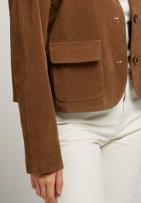 Veste en daim marron avec un col haut, des boutons à l'avant, deux poches poitrine et des manches larges, portée sur un pantalon blanc. Texture douce et design ajusté.