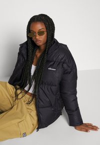 Black puffer jacket with a quilted design, featuring a logo on the left chest, paired with beige baggy pants and green tinted sunglasses.