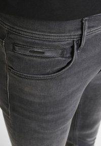 Tamnosive traperice od denima klasičnog dizajna, s stražnjim džepom koji ima pravokutnu aplikaciju i suptilne detalje šivanja.