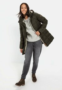 Olivengrøn pufferjakke med hætte, lynlåslukning og quiltet design. Bæres over en lys grå fleece-trøje og blå jeans, parret med brune støvler.