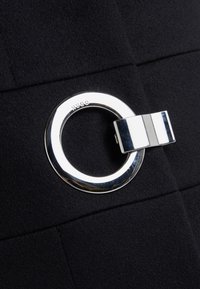 Tela negra con una textura suave que presenta un detalle de anillo y cierre plateados. El anillo está grabado con "HUGO."