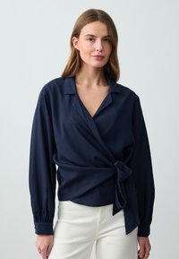 Camicia avvolgente blu navy con maniche lunghe, dotata di colletto e dettaglio con nodo laterale. Realizzata in tessuto liscio, abbinata a pantaloni chiari.