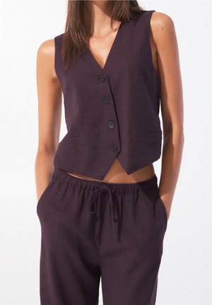 Femme portant un gilet sans manches violet foncé à boutons et un pantalon assorti avec cordon, les mains dans les poches, sur fond blanc.
