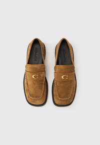 Καφέ σουέντ μοκασίνια με στρογγυλεμένη μύτη, χρυσές μεταλλικές λεπτομέρειες και αντίθετη μαύρη σόλα. Διαθέτουν ορατές ραφές.