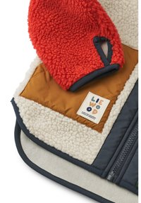 Kinderjacke mit einem flauschigen roten Fleece-Ärmel, sandfarbenen und cremefarbenen Panele und einem navyblauen Saum. Ein Etikett mit 'LIEWOOD' ist sichtbar.