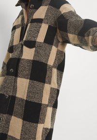 Skjortjacka i ullblandning med ett svart och beige rutmönster, klädd framknäppning och två bröstfickor. Mjuk textur.