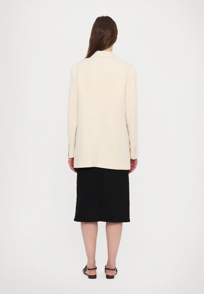 Blazer beige su misura con maniche lunghe, dettaglio della cucitura posteriore, abbinato a una gonna nera al ginocchio e sandali neri. Design minimalista.
