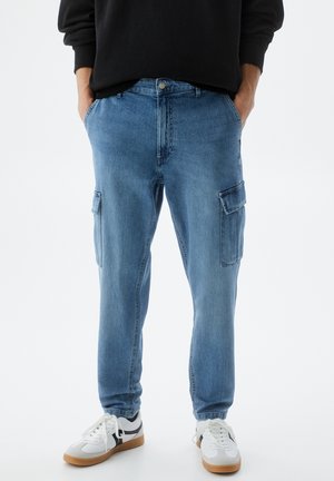 Homme portant un pantalon cargo en denim bleu avec poches latérales, sweat-shirt noir, baskets blanches, debout les mains dans les poches sur fond blanc.