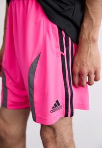 Rožinės sportinės šortai su juodais ir pilkais akcentais, turintys šonines juosteles, lygią tekstūrą ir "Adidas" logotipą ant apačios.