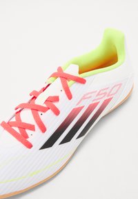 Scarpa da ginnastica bianca con lacci rosa, colletto giallo neon e strisce nere. Presenta una parte superiore testurizzata e una suola in gomma.