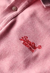 Czerwony haftowany logo na jasnoróżowej koszulce polo, charakteryzująca się teksturowanym materiałem i placketem na dwa guziki z subtelnym kołnierzykiem.