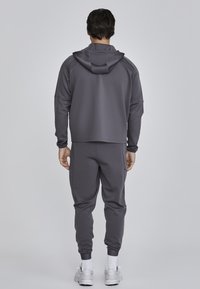 Pánsky atletický outfit v tmavošedej farbe. Obsahuje mikinu s kapucňou a zúžené tepláky s rebrovanými manžetami. Vyrobené z hladkej, textúrovanej tkaniny.