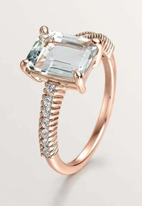 Anillo de oro rosa con una gran piedra preciosa en corte esmeralda, rodeada de pequeñas piedras blancas, con un diseño de banda retorcida y acabado suave.
