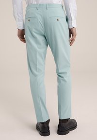 Mint green tailored trousers with a slim fit, back pockets, and brass button accents. Worn with black shoes and a white long-sleeve shirt.