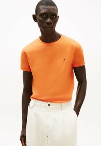 T-shirt arancione a maniche corte in cotone, con un piccolo logo ricamato sul lato sinistro del petto, abbinato a pantaloni bianchi su misura.