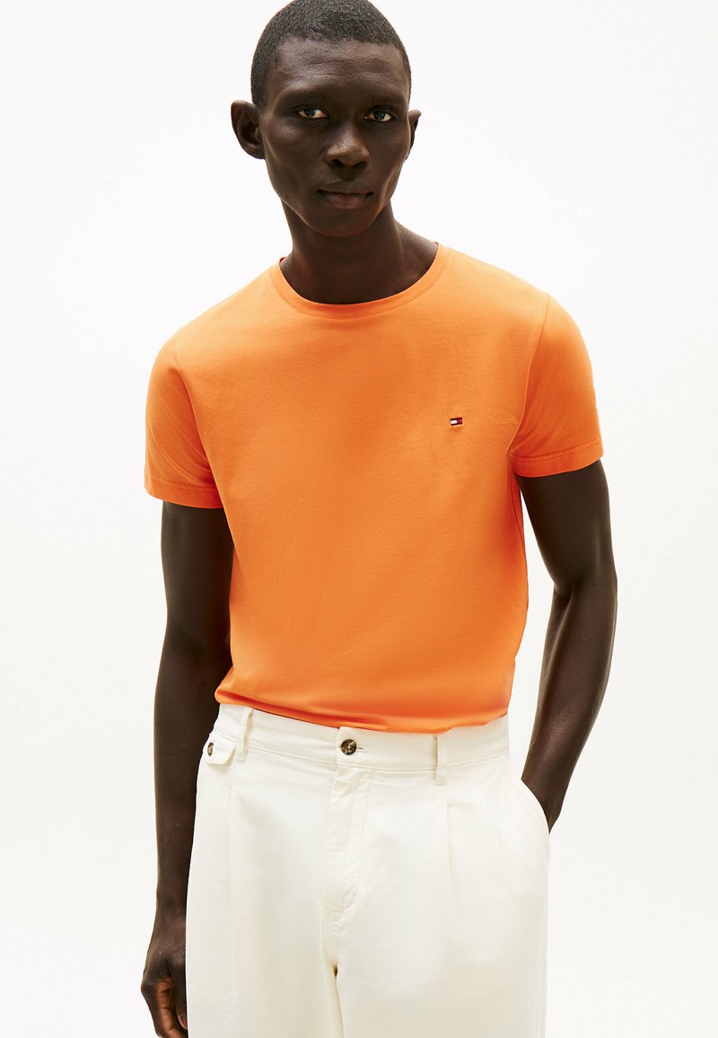 T-shirt arancione a maniche corte in cotone, con un piccolo logo ricamato sul lato sinistro del petto, abbinato a pantaloni bianchi su misura.