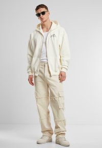 Hanorac crem cu fermoar și buzunare tip cangur, purtat peste un top alb fără mâneci; asortat cu pantaloni cargo deschiși la culoare și pantofi sport bej.