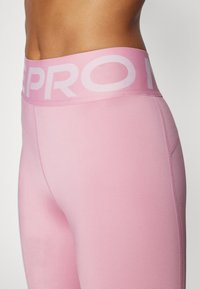 Leggings rosa con un ampio cinturino che presenta un marchio bianco. Texture liscia, design aderente e cuciture laterali eleganti.