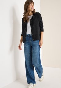 Schwarze Strickjacke mit Knöpfen, darunter ein weißes T-Shirt, hellblaue weite Jeans und weiße Sneaker mit schwarzen Akzenten.