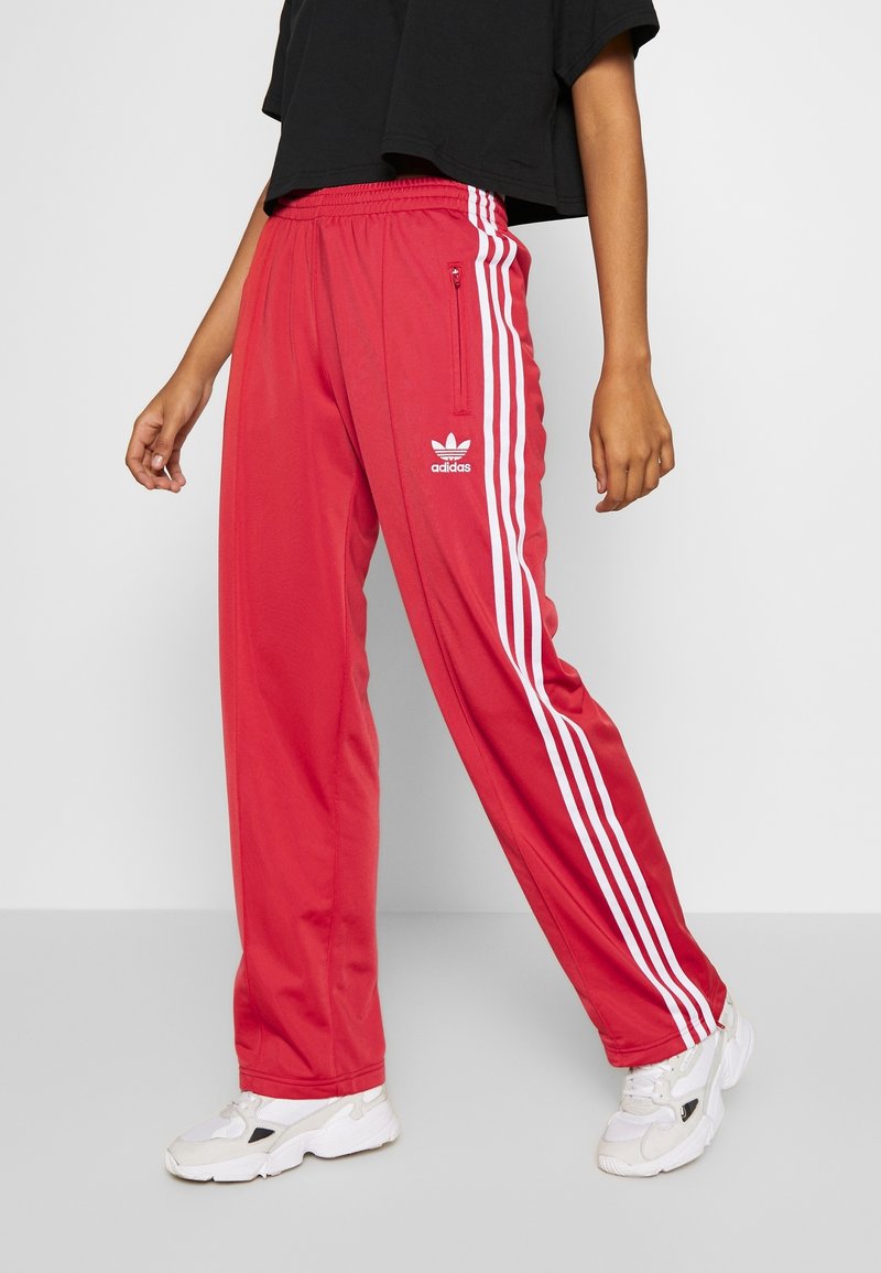 Pantalons de sport rouges en matériau léger avec des rayures blanches sur les côtés et un logo brodé. Taille élastique avec détails de poche zippée.