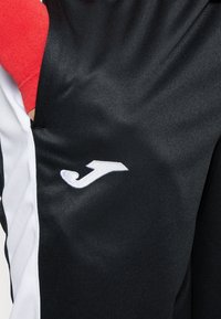 Czarne sportowe spodenki z białymi bocznymi paskami, z białym haftowanym logo "J" po lewej stronie. Gładka, lekka tkanina.