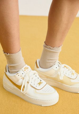 Fødder iført hvide og gyldne Nike Air Force 1 sneakers med beige ribstrikkede sokker på gul gulv.