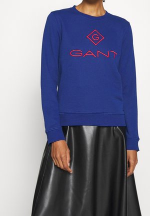 Blå sweatshirt med rund halsringning, prydd med en röd broderad logotyp och texten "GANT" på framsidan. Buren över en svart läderkjol.