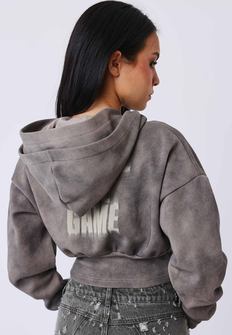 Femme aux longs cheveux noirs portant un sweat à capuche gris court avec le texte "GAME" au dos et un jean gris déchiré taille haute, vue de profil.