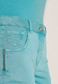 Nahaufnahme von türkisfarbenen Hosen mit einem Reißverschlusstaschen und einem Metallknopf, kombiniert mit einem passenden türkisfarbenen Oberteil.