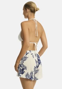 Een vrouw met blond haar dat in een knot is gebonden, draagt een witte bikinitop en bloemenpatronen shorts, poseert met haar rug naar de camera.