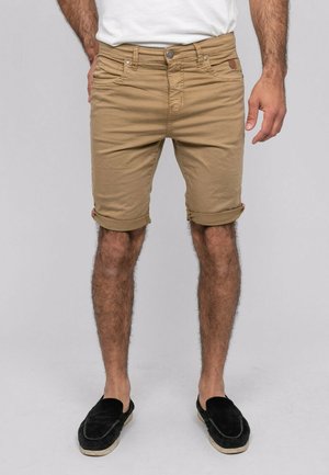 Homme portant un short beige roulé, des chaussures noires sans lacets et un t-shirt blanc, debout devant un fond uni.