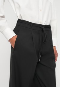 Mustad püksid nööriga vöökohaga, voldid ja küljetaskud. Valmistatud pehmest kangast, millel on sile tekstuur. Sobitatud valge nööbitava särgiga.