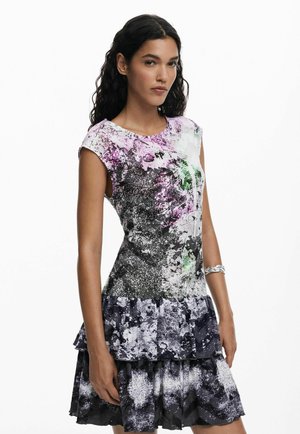 Donna con lunghi capelli ricci che indossa un vestito aderente senza maniche, con motivi astratti in viola, nero e verde, in piedi con una mano rilassata.