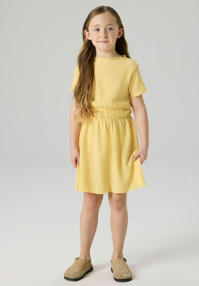 Jeune fille aux longs cheveux portant une robe jaune texturée et des chaussures beige à enfiler, debout devant un fond clair uni.