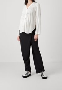 Vit långärmad blus med veck, kombinerad med lösa svarta byxor och glänsande silvriga slip-on skor, som framhäver tygtexturer och färger.